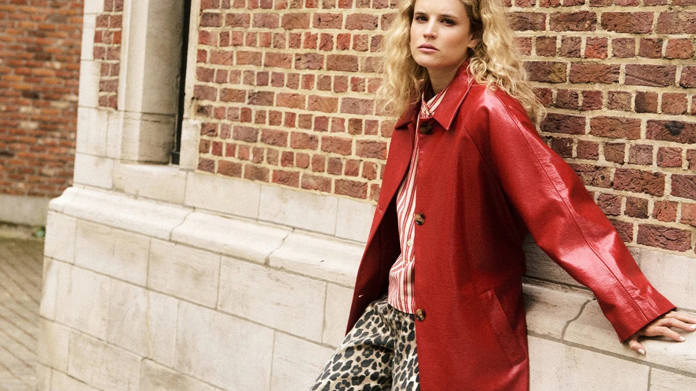 Woman in a red coat and leopard print pants leaning against a brick wall.
